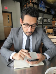 A man in a business suit is sitting at a table in a modern cafe or restaurant. He is looking intently at a smartphone, holding a pen, and has a notebook open on the table in front of him. The setting includes wooden chairs, and there are monochrome photographs on the wall and televisions mounted in the background.