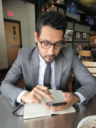 A man in a business suit is sitting at a table in a modern cafe or restaurant. He is looking intently at a smartphone, holding a pen, and has a notebook open on the table in front of him. The setting includes wooden chairs, and there are monochrome photographs on the wall and televisions mounted in the background.