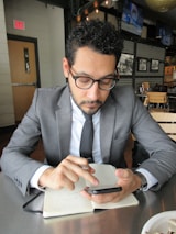 A man in a business suit is sitting at a table in a modern cafe or restaurant. He is looking intently at a smartphone, holding a pen, and has a notebook open on the table in front of him. The setting includes wooden chairs, and there are monochrome photographs on the wall and televisions mounted in the background.