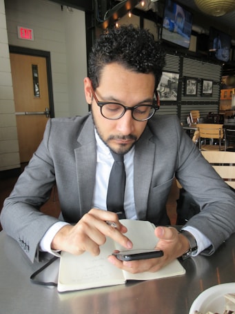 A man in a business suit is sitting at a table in a modern cafe or restaurant. He is looking intently at a smartphone, holding a pen, and has a notebook open on the table in front of him. The setting includes wooden chairs, and there are monochrome photographs on the wall and televisions mounted in the background.