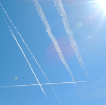Several contrails intersect against a clear blue sky, casting linear white streaks. The sun is visible in the top right corner, creating a lens flare effect.