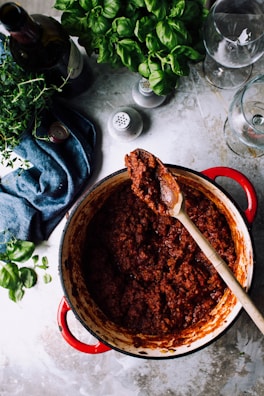 A red pot filled with rich, thick tomato sauce sits on a marble countertop. A wooden spoon is resting on the edge of the pot with sauce on it. Surrounding the pot are fresh green basil leaves, a bottle of wine, two empty wine glasses, and a blue cloth. The setting appears to be a kitchen preparing for a meal.