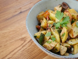 A bowl filled with roasted potato pieces garnished with fresh parsley leaves. The potatoes are browned and seasoned, mixed with bits of vegetables and herbs. The bowl is placed on a wooden surface.