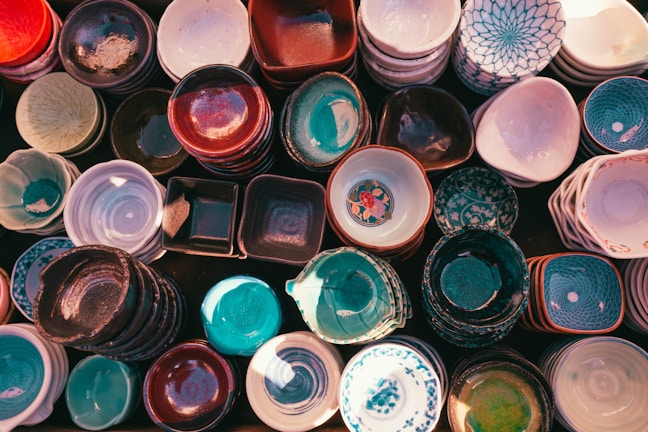 An assortment of ceramic bowls and dishes is arranged closely together. The variety includes different colors, patterns, and shapes, with some featuring intricate designs and others having solid colors. The light casts soft, warm highlights on the surfaces, creating a cozy and inviting look.