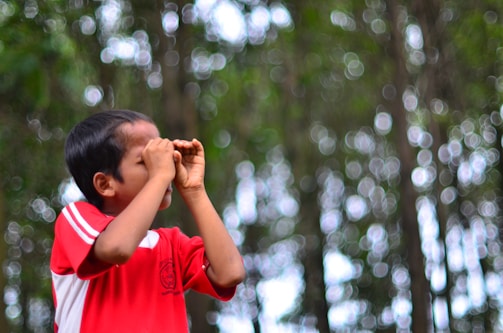 A young child wearing a red shirt appears to be looking through imaginary binoculars made with their hands. The background consists of a bokeh effect with bright circles of light among blurred green trees.