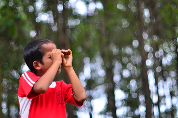 A young child wearing a red shirt appears to be looking through imaginary binoculars made with their hands. The background consists of a bokeh effect with bright circles of light among blurred green trees.