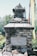 A stone monument constructed from stacked rocks, with an inscription dedicated to mountaineers who lost their lives. The background features some greenery and colorful vertical flags or banners, likely indicative of a cultural or memorial site.