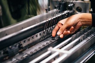 A hand is carefully operating a complex piece of machinery, involving several metal rods and strings, likely part of a textile manufacturing process.