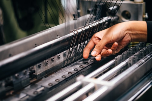 A hand is carefully operating a complex piece of machinery, involving several metal rods and strings, likely part of a textile manufacturing process.
