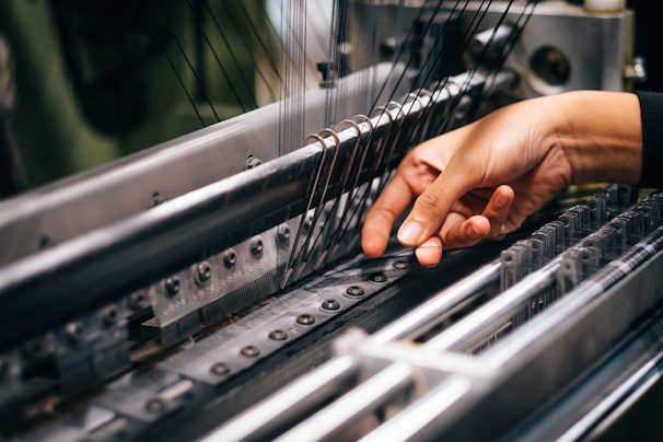 A hand is carefully operating a complex piece of machinery, involving several metal rods and strings, likely part of a textile manufacturing process.
