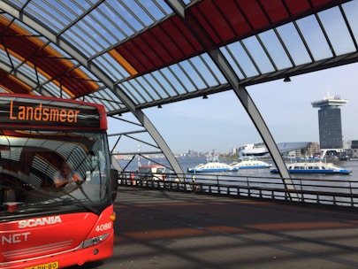 A bright red Scania bus labeled 'Landsmeer' is parked in a modern, open-air transit terminal with a geometric roof featuring red and yellow panels. In the background, a river with white and blue ferries is visible, along with a tall, modern building and other city structures.