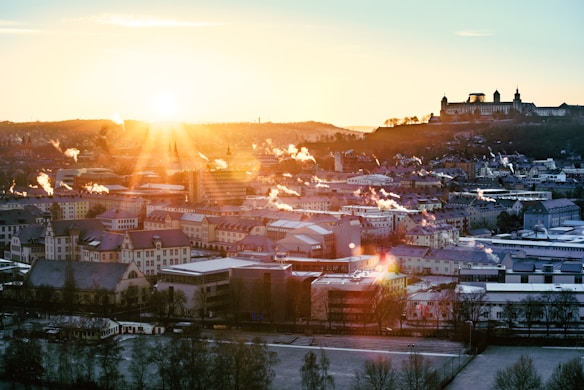 A cityscape during sunrise with sunlight illuminating the scene, casting a warm glow over the buildings. Smoke rises from various points in the city, adding to the atmospheric setting. In the background, a prominent structure with towers is visible on a hill.