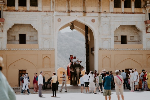 A bustling scene unfolds at the entrance of an ornate historical structure with an arched doorway. Tourists gather around, some taking photos while others converse or walk by. An elephant with two riders is making its way through the entry, adding a sense of cultural richness and tradition. The architecture features intricate patterns and neutral tones, accentuating the historical significance of the location.