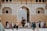 A bustling scene unfolds at the entrance of an ornate historical structure with an arched doorway. Tourists gather around, some taking photos while others converse or walk by. An elephant with two riders is making its way through the entry, adding a sense of cultural richness and tradition. The architecture features intricate patterns and neutral tones, accentuating the historical significance of the location.