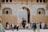 A bustling scene unfolds at the entrance of an ornate historical structure with an arched doorway. Tourists gather around, some taking photos while others converse or walk by. An elephant with two riders is making its way through the entry, adding a sense of cultural richness and tradition. The architecture features intricate patterns and neutral tones, accentuating the historical significance of the location.