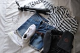 A neatly arranged set of clothing and accessories on a bed includes a striped long-sleeve shirt, denim shorts, white athletic shoes, a black backpack, a camera tripod, and a pair of eyeglasses.