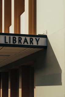 An architectural structure with a sign reading 'LIBRARY' in bold, dark letters. The building features tall, narrow windows and cast shadows on a light-colored wall, evoking a minimalist aesthetic.