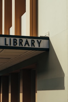 An architectural structure with a sign reading 'LIBRARY' in bold, dark letters. The building features tall, narrow windows and cast shadows on a light-colored wall, evoking a minimalist aesthetic.