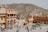 A large, historical fort complex with sandy-colored walls is populated by tourists and an elephant. It is situated in an arid, hilly landscape, providing a backdrop of rocky hillsides. The architecture features arched doorways and intricate balconies.