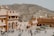A large, historical fort complex with sandy-colored walls is populated by tourists and an elephant. It is situated in an arid, hilly landscape, providing a backdrop of rocky hillsides. The architecture features arched doorways and intricate balconies.