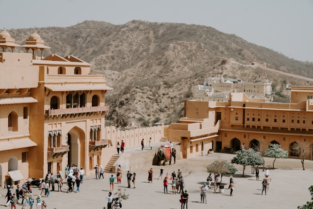 A large, historical fort complex with sandy-colored walls is populated by tourists and an elephant. It is situated in an arid, hilly landscape, providing a backdrop of rocky hillsides. The architecture features arched doorways and intricate balconies.
