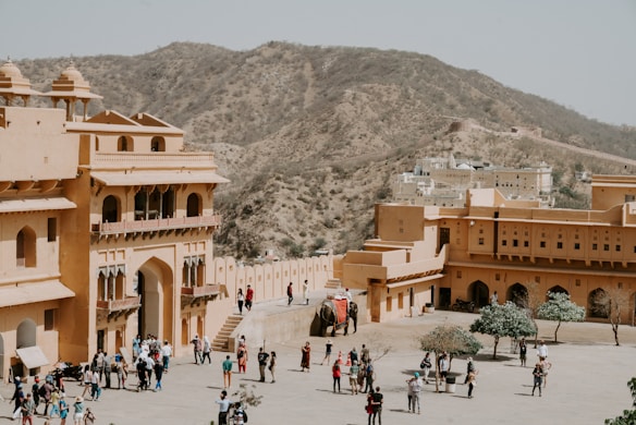 A large, historical fort complex with sandy-colored walls is populated by tourists and an elephant. It is situated in an arid, hilly landscape, providing a backdrop of rocky hillsides. The architecture features arched doorways and intricate balconies.