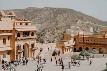 A large, historical fort complex with sandy-colored walls is populated by tourists and an elephant. It is situated in an arid, hilly landscape, providing a backdrop of rocky hillsides. The architecture features arched doorways and intricate balconies.