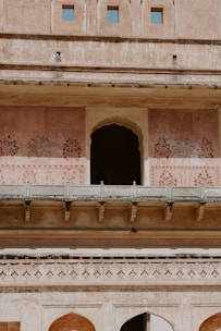 An ornate architectural facade featuring arched doorways and intricate painted patterns on a pinkish wall. The structure is multi-leveled with a series of small, square windows at the top. The architecture reflects historical design with decorative floral motifs.