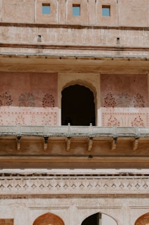 An ornate architectural facade featuring arched doorways and intricate painted patterns on a pinkish wall. The structure is multi-leveled with a series of small, square windows at the top. The architecture reflects historical design with decorative floral motifs.