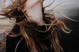 Long, flowing hair is captured in motion against a blurred background. The strands of hair have a natural mix of colors ranging from dark brown to lighter blond shades. The motion suggests a dynamic and free-spirited moment.