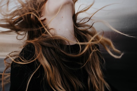 Long, flowing hair is captured in motion against a blurred background. The strands of hair have a natural mix of colors ranging from dark brown to lighter blond shades. The motion suggests a dynamic and free-spirited moment.