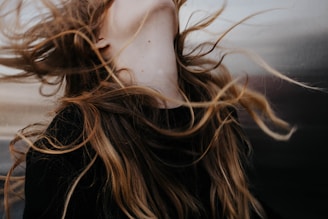 Long, flowing hair is captured in motion against a blurred background. The strands of hair have a natural mix of colors ranging from dark brown to lighter blond shades. The motion suggests a dynamic and free-spirited moment.