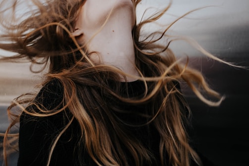 Long, flowing hair is captured in motion against a blurred background. The strands of hair have a natural mix of colors ranging from dark brown to lighter blond shades. The motion suggests a dynamic and free-spirited moment.