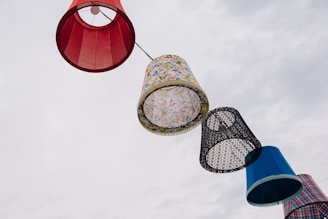 A series of large, colorful lampshades are strung together, suspended against a cloudy sky. The lampshades vary in patterns and colors, including red, floral, black and white lace, and a blue plaid design.
