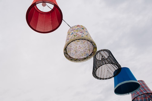 A series of large, colorful lampshades are strung together, suspended against a cloudy sky. The lampshades vary in patterns and colors, including red, floral, black and white lace, and a blue plaid design.