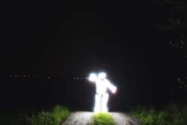 A brightly illuminated figure stands on a narrow path surrounded by darkness. The intense glow obscures details, creating a mysterious and surreal effect. Sparse vegetation lines the path, with faint points of light visible in the distant background.