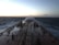 A large ship navigates through rough seas with waves crashing over its deck. The sky is overcast with hints of light suggesting early morning or late evening. Long pipelines run along the length of the ship, which appears sturdy and industrial.