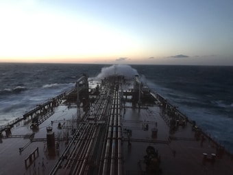 A large ship navigates through rough seas with waves crashing over its deck. The sky is overcast with hints of light suggesting early morning or late evening. Long pipelines run along the length of the ship, which appears sturdy and industrial.