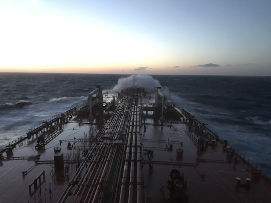 A large ship navigates through rough seas with waves crashing over its deck. The sky is overcast with hints of light suggesting early morning or late evening. Long pipelines run along the length of the ship, which appears sturdy and industrial.