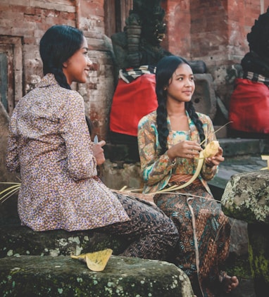 woman holding woven coconut leaf beside woman sitting while holding knife