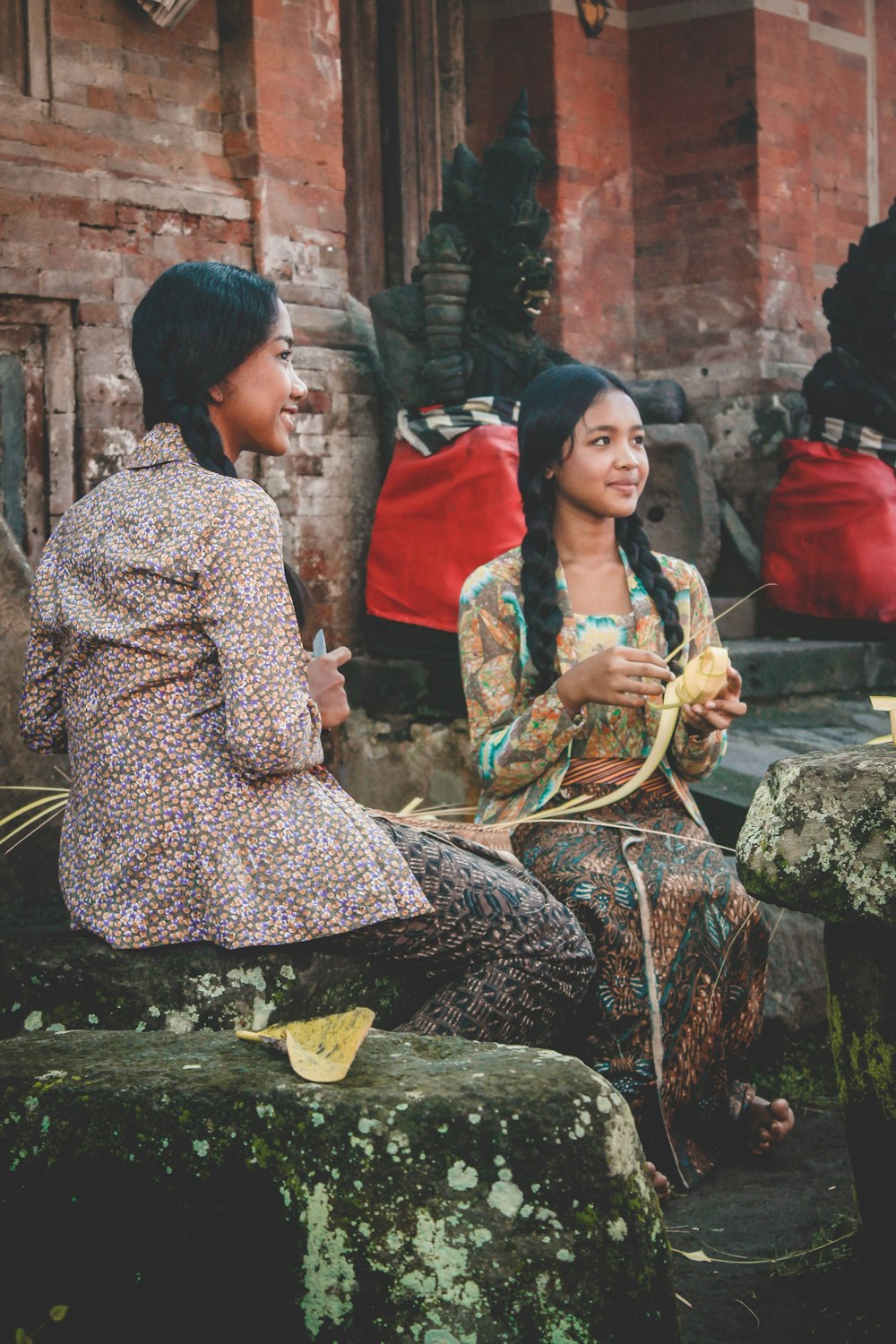 ナイフを持ちながら座っている女性の横で編んだココナッツの葉を持っている女性