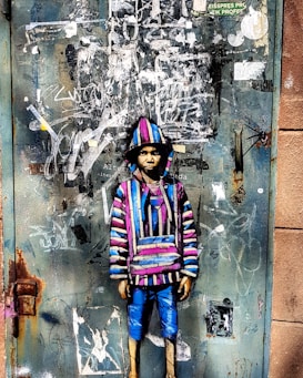 A graffiti artwork on a weathered metal door featuring a figure in a colorful striped hoodie and blue pants. The door is covered with layers of peeling paint and scribbled graffiti, creating a textured and urban backdrop.