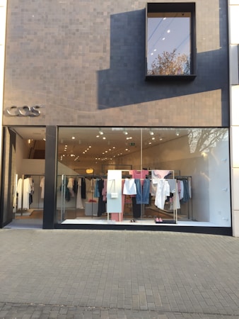 A modern fashion retail store with a minimalist facade. The storefront features a large glass window displaying a clothing rack with various garments, including shirts, pants, and dresses in pastel and neutral colors. The exterior wall is covered in dark gray tiles, with a small, rectangular window reflecting a tree.