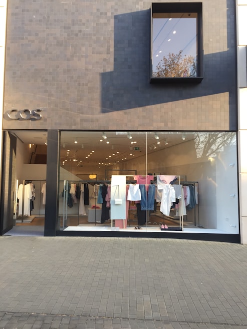 A modern fashion retail store with a minimalist facade. The storefront features a large glass window displaying a clothing rack with various garments, including shirts, pants, and dresses in pastel and neutral colors. The exterior wall is covered in dark gray tiles, with a small, rectangular window reflecting a tree.