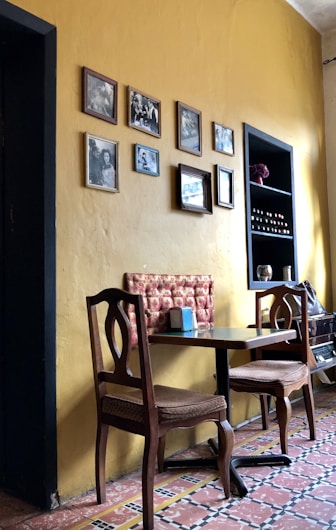 A cozy corner of a room with a small table and two wooden chairs, accompanied by a cushioned bench against a yellow wall. The wall is decorated with several framed black and white photographs. A shelf next to the table holds various small objects, including bottles and a red decorative piece. The floor features a patterned tile design.