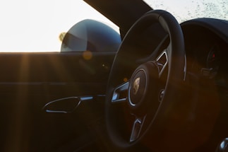 An interior view of a car showcasing the steering wheel with a visible logo, alongside the door handle. Sunlight is streaming through the window, creating lens flares, while the background features a frosted window.