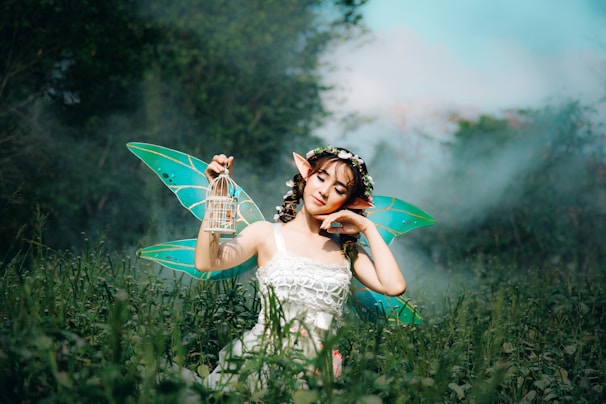 A woman dressed as a fairy with translucent blue-green wings stands in a lush, green forest. She is wearing a white dress and has a flower crown on her head. She holds a small birdcage in one hand and poses gracefully with her eyes closed. Mist surrounds the scene, adding a mystical atmosphere.