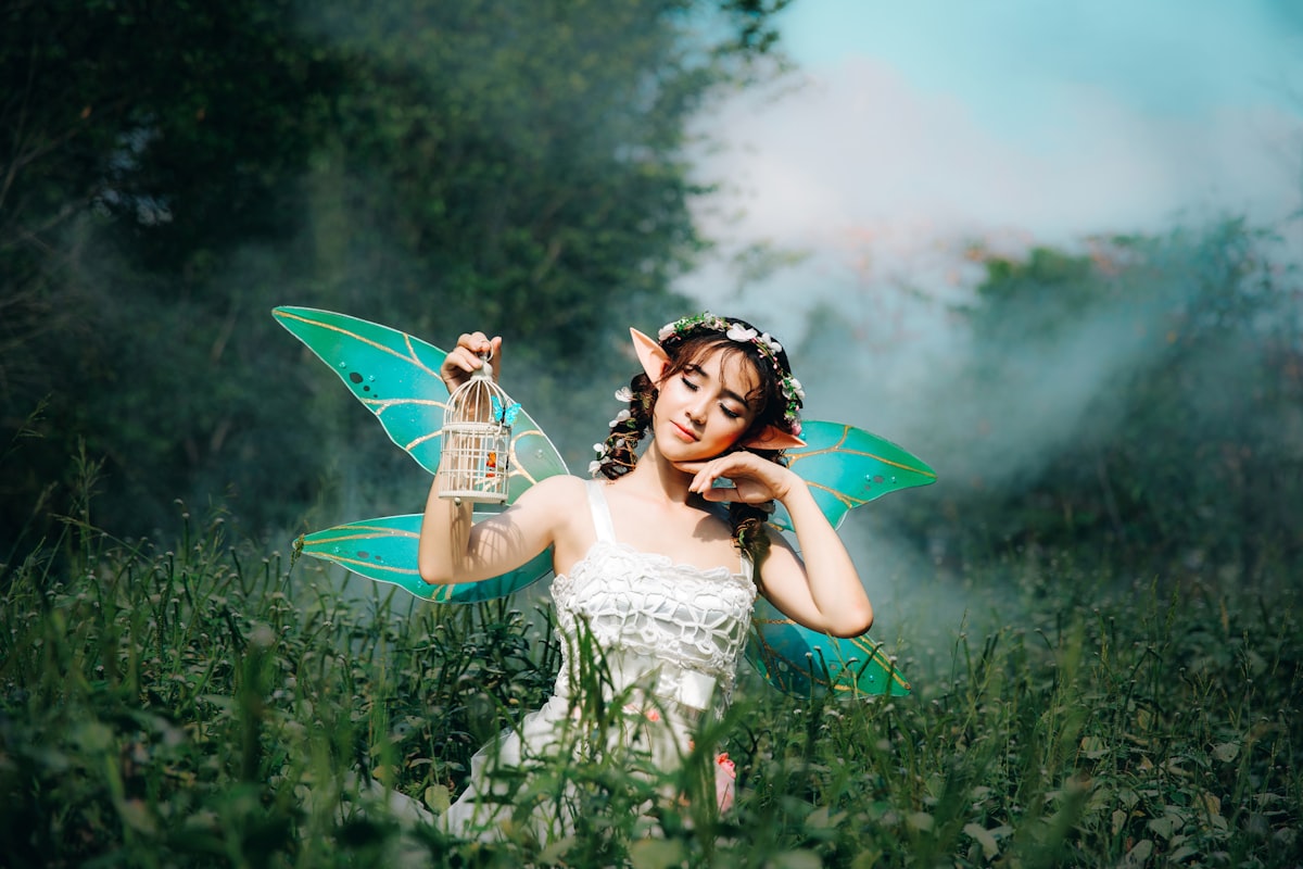 a light skinned fairy with huge ears and bright turquoise wings in a misty field of grass.  She has white flowers in her hair and is wearing a white dress and holds a tiny thie bird cage.