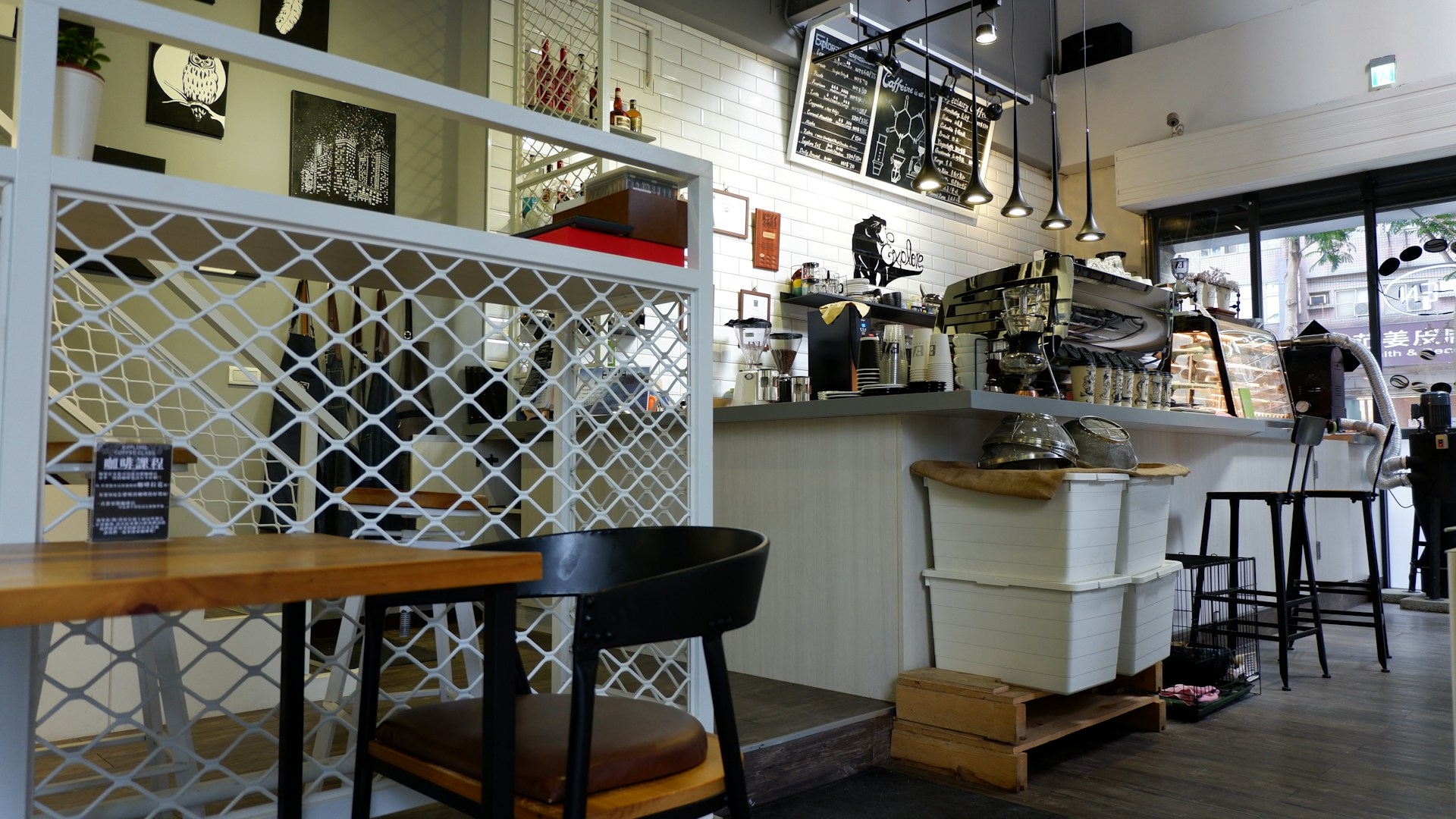 A cozy café interior with a modern, clean design. The setting includes wooden chairs and tables, a counter with various coffee-making equipment, and a menu board displayed above. Decorations include paintings on the walls, minimalist lighting fixtures, and a display case with pastries. The flooring and partitions feature wood and metal elements, contributing to a relaxed atmosphere.