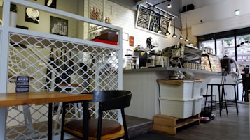 A cozy caf&eacute; interior with a modern, clean design. The setting includes wooden chairs and tables, a counter with various coffee-making equipment, and a menu board displayed above. Decorations include paintings on the walls, minimalist lighting fixtures, and a display case with pastries. The flooring and partitions feature wood and metal elements, contributing to a relaxed atmosphere.
